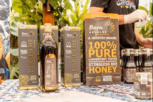Bayu Kelulut honey products including jars, bottles, and boxes on a table with a person in the background.